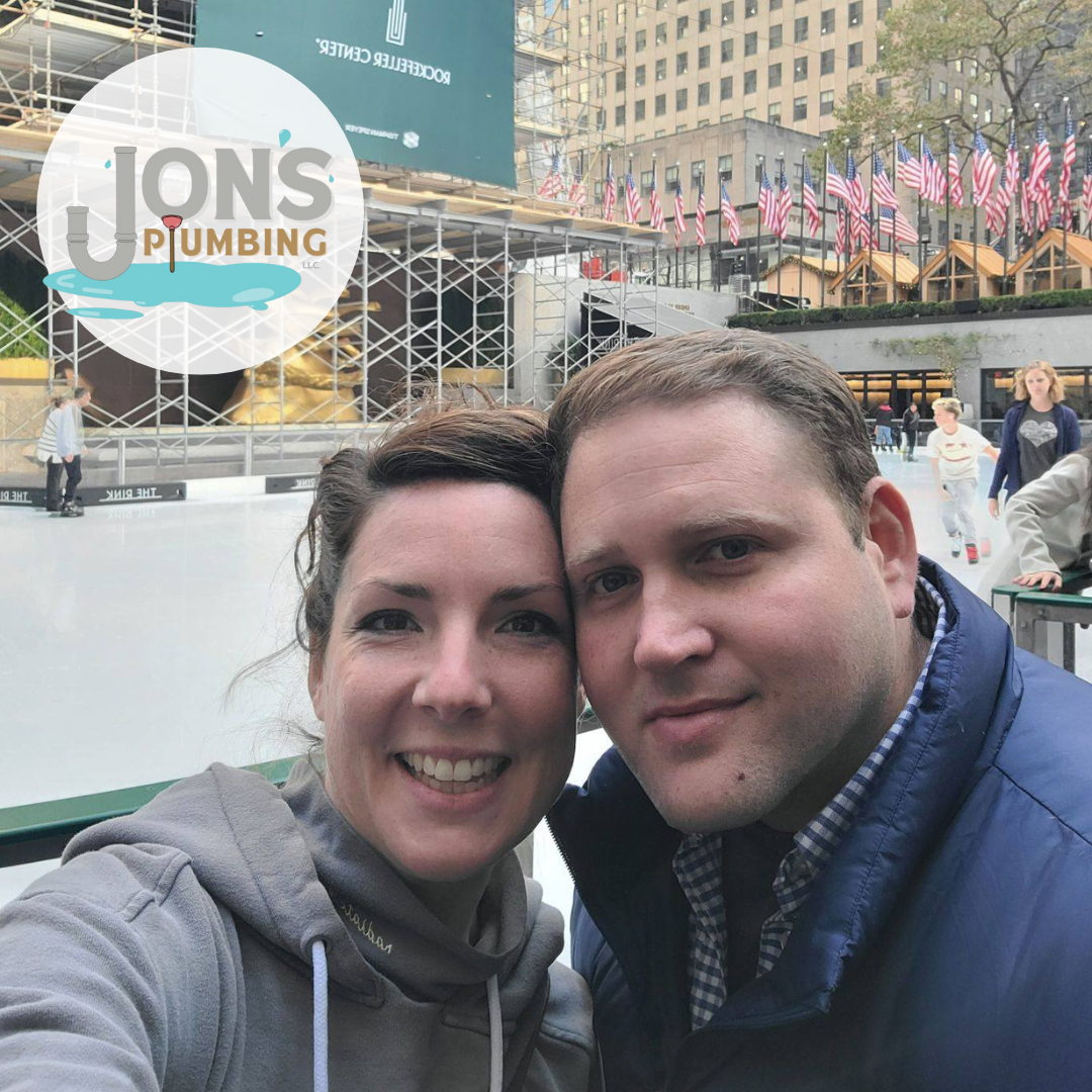 Jon from Jon’s Plumbing and his wife smiling together at the Rockefeller Center ice rink in New York City, with American flags and scaffolding in the background. The Jon’s Plumbing logo is overlaid in the corner.